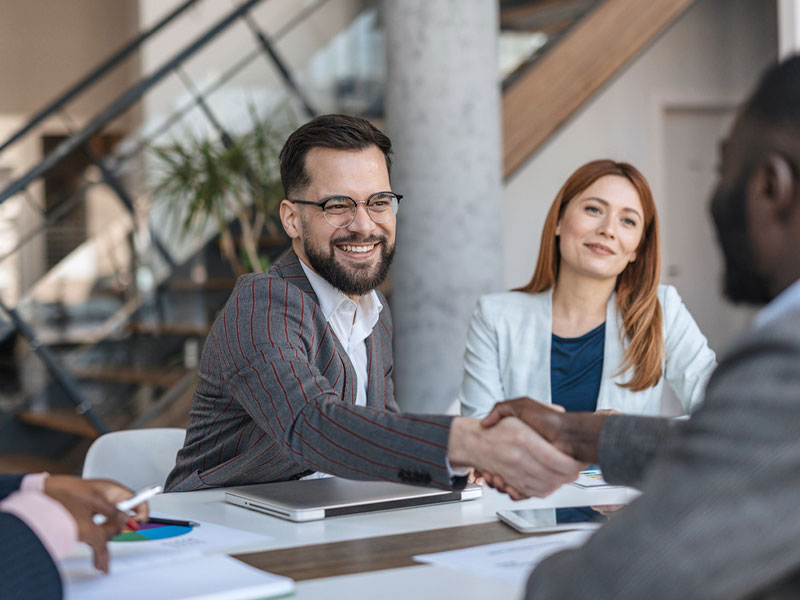 A smiling man in glasses shakes hands with another person at a business meeting.