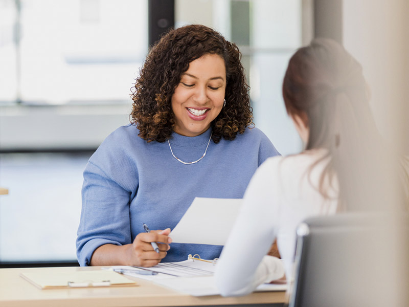 A woman in a blue sweater smiling while reviewing documents in an office setting.