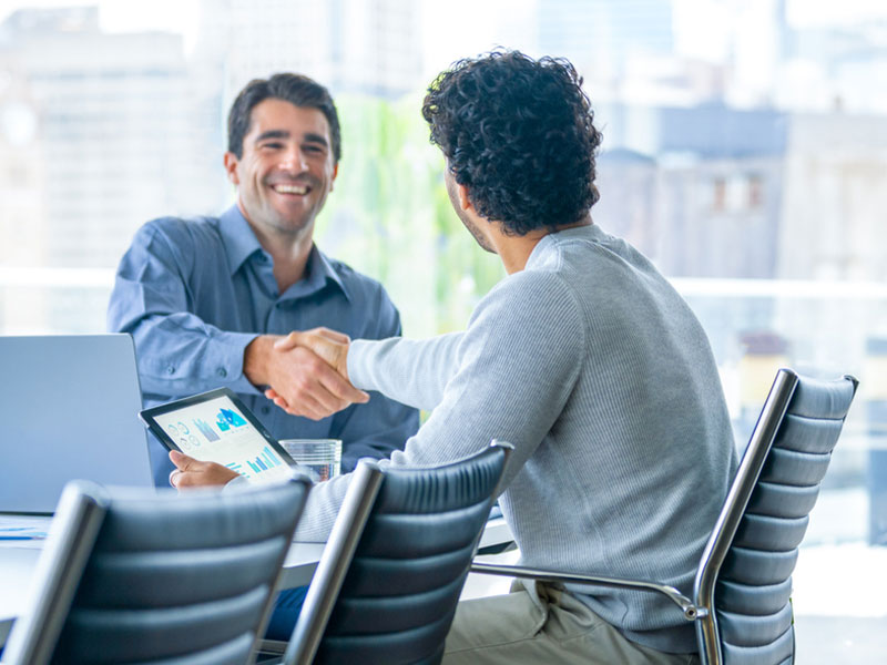 Two men shaking hands in a modern office setting with a tablet on the table.