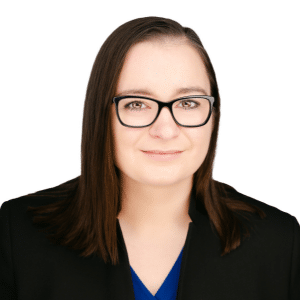 A woman with glasses and straight brown hair, wearing a black jacket, smiling against a white background.