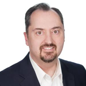 A person with short dark hair and a goatee, wearing a white shirt and dark blazer, smiling at the camera.