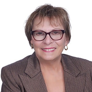 A woman wearing glasses and a brown blazer smiles against a white background.